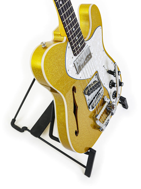 A golden Txxx Custom Thinline '72 Partscaster with B5 Bigsby rests on a stand, highlighted by a simple white background.