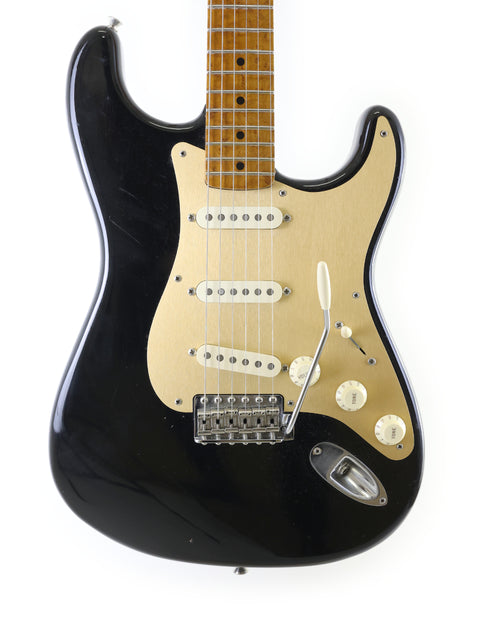 A black Fender AVRI ‘56 Stratocaster electric guitar featuring a striking gold pickguard, showcasing its classic design.