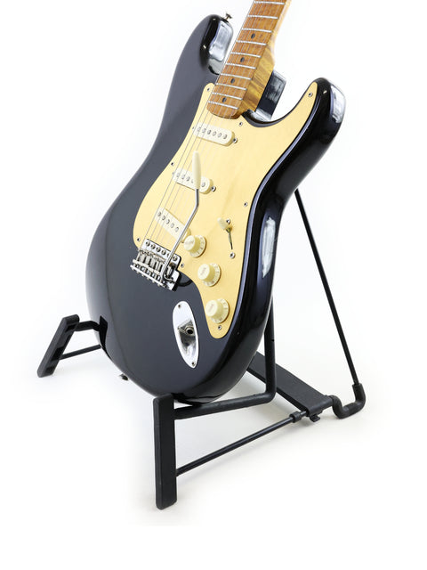 A black Fender AVRI ‘56 Stratocaster guitar displayed on a stand, set against a bright white background for emphasis.