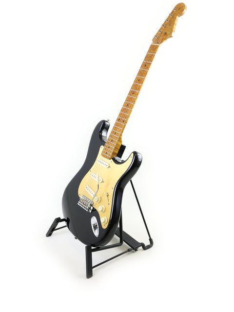 A black Fender AVRI ‘56 Stratocaster guitar elegantly displayed on a stand, set against a bright white backdrop.