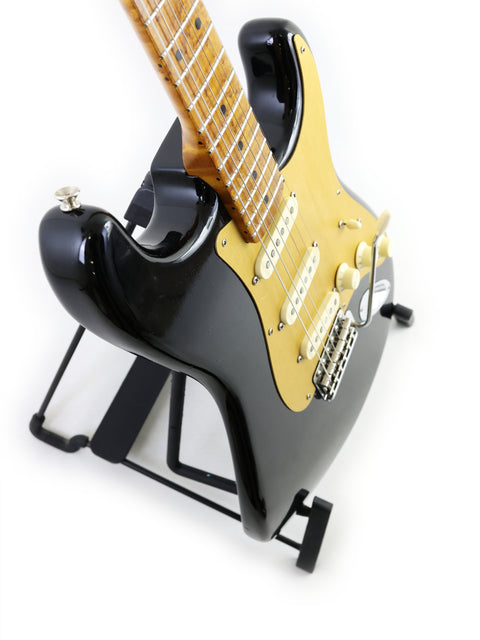 A black and gold Fender AVRI ‘56 Stratocaster guitar on a stand, emphasizing its sleek design and craftsmanship.