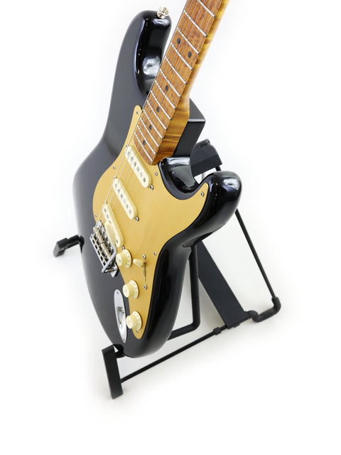 A black guitar, the Fender AVRI ‘56 Stratocaster, elegantly positioned on a stand with a stark white backdrop.