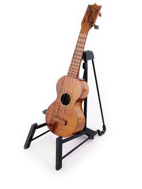 A historic Kumalae Style 2 ukulele, crafted in Hawaii circa 1913, displayed on a sleek black stand, highlighting its beauty.