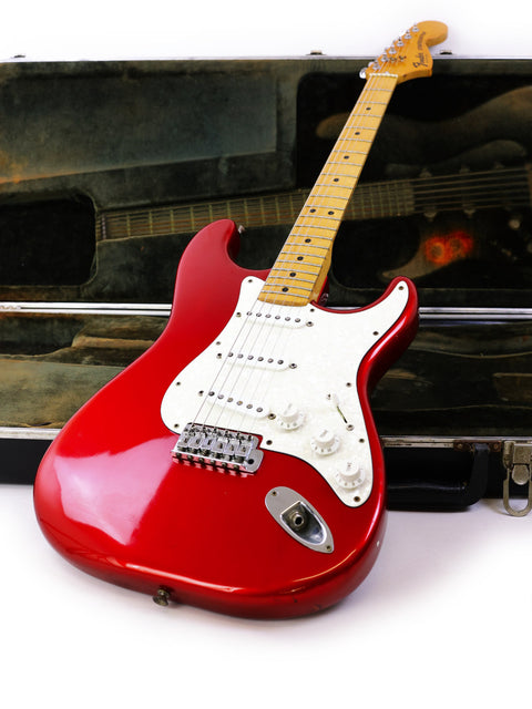 A red 1979 Fender Stratocaster guitar nestled in a case, with a guitar pick resting beside it, highlighting its classic style.
