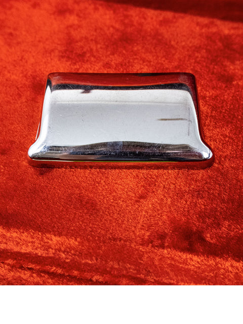 A silver metal component of a 1964 Fender L Series Stratocaster in Fiesta Red displayed on an orange velvet background.