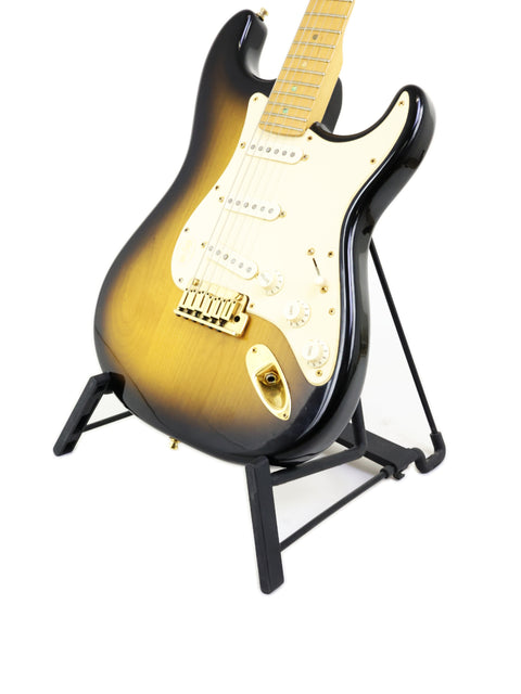 A sunburst electric guitar with a maple neck on a black stand against a white background.
