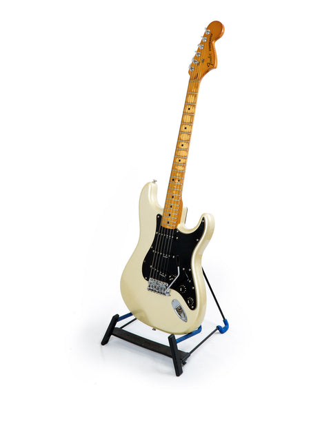 A white USA-made Fender 25th Anniversary Stratocaster guitar displayed on a stand, showcasing its black pick guard.