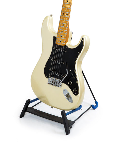 A white guitar on a stand, the Fender 25th Anniversary Stratocaster from 1979, highlighted by a contrasting black pick guard.