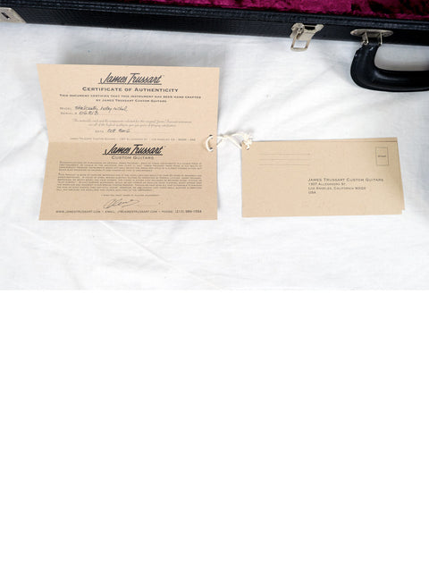 An open black and red suitcase displays a note and envelope, with a James Trussart Holey Nickel Steelcaster guitar nearby.