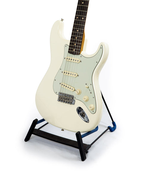 white guitar, the Fender '60s Strat Partscaster, rests on a black base stand, emphasizing its stylish design and quality.