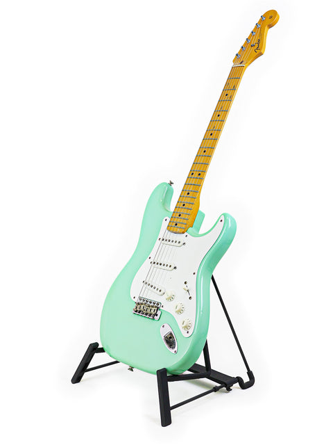 A classic green Fender AVRI ‘57 Stratocaster guitar displayed on a stand with a bright white backdrop.