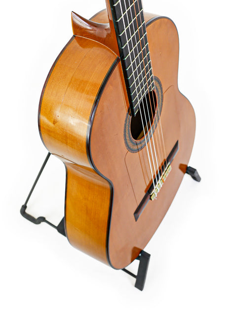 A guitar resting on a stand in front of a plain white background, emphasizing its craftsmanship. M.G. Contreras Calle Mayor 80 - Spain 1973.