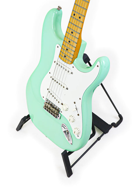 A vintage 1985 green Fender AVRI ‘57 Stratocaster electric guitar on a stand, highlighting its iconic style.