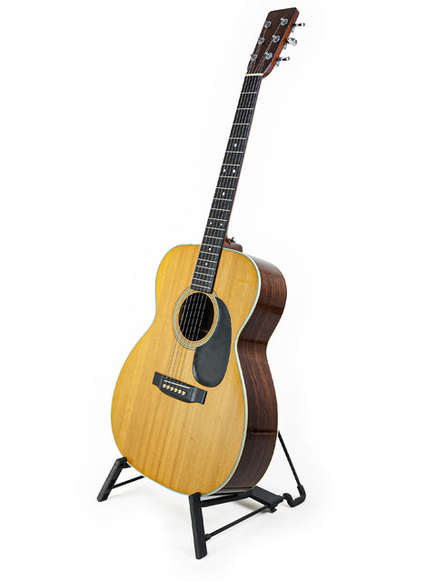 A vintage Martin 000-28 guitar from 1976 resting on a stand, set against a stark white background.

