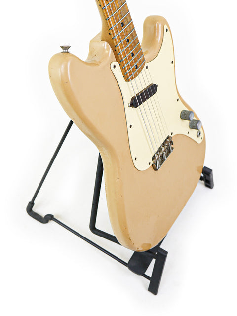 A vintage tan Fender Musicmaster guitar from 1957 on a stand, set against a minimalist white background for a striking effect.