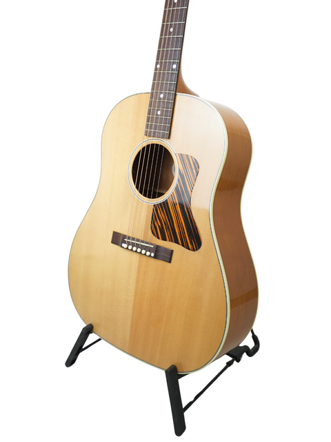 Displaying a 2013 Gibson J-35 guitar on a stand, this image highlights its beauty against a simple white background.