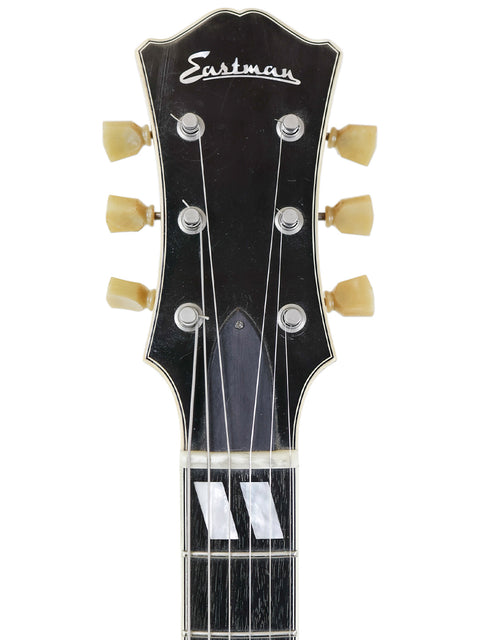 Headstock of an Eastman guitar with tuning pegs and strings on a white background.