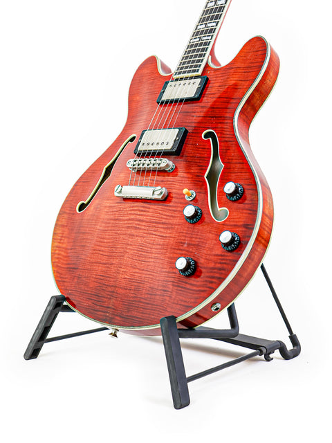 Red semi-hollow electric guitar on a black stand against a white background.