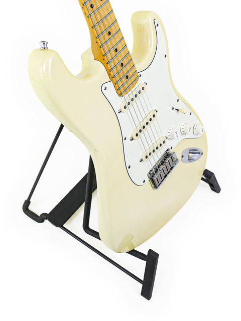 A cream-colored electric guitar on a black stand against a white background.