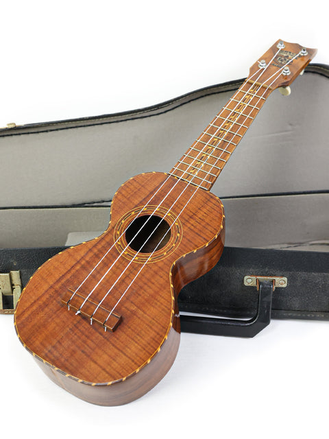 white background features a 1920s Kumalae ukulele securely placed in its case.
