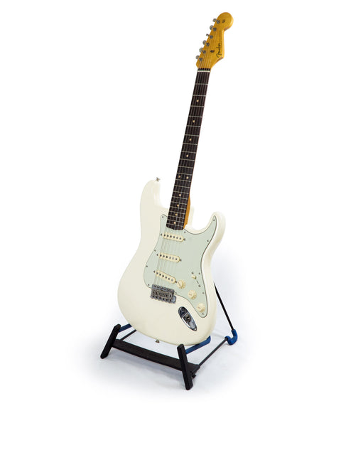A Fender '60s Strat Partscaster in Olympic White rests on a black stand, emphasizing its sleek design and craftsmanship.

