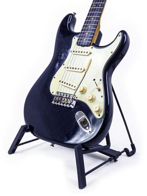 A black 1963 Fender L-Series Stratocaster guitar elegantly positioned on a stand with a stark white background.