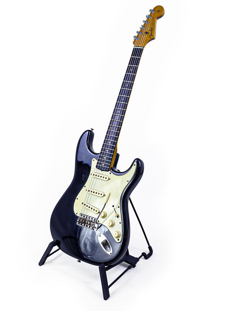 A black and white image of a 1963 Fender L-Series Stratocaster guitar elegantly displayed on a stand.
