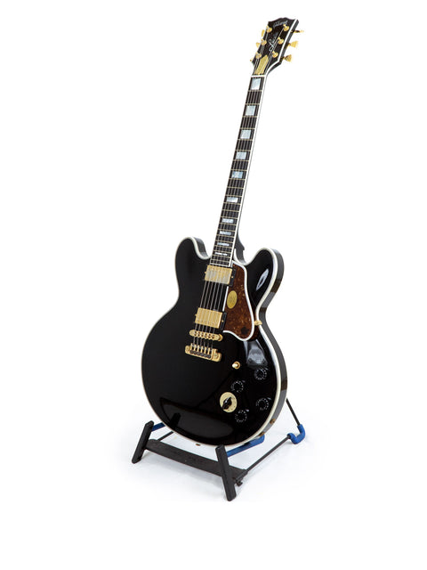 A black guitar, the Gibson Lucille, displayed on a stand with a white backdrop, featuring signatures from BB King and Eric Clapton.