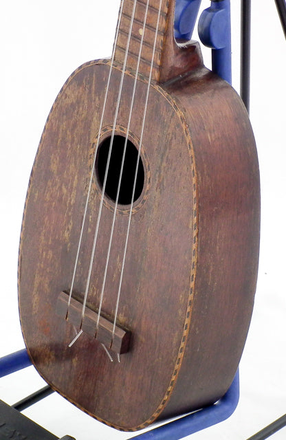 A blue stand holds a vintage Kamaka Pineapple Ukulele from 1930, emphasizing its historical significance and beauty.