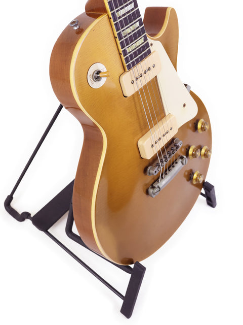 A gold guitar, the Gibson Les Paul 1956 Reissue, elegantly positioned on a stand against a bright white background.
