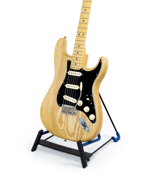 A natural oiled ash Fender Stratocaster guitar from 2015, resting on a sleek black stand, highlighting its craftsmanship.
