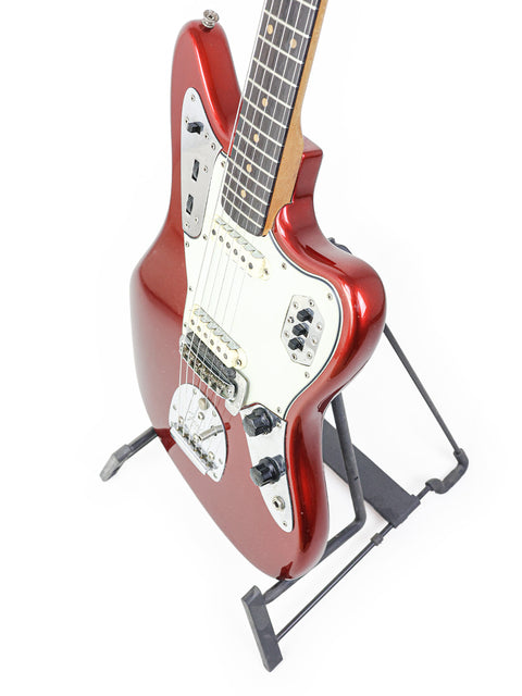 A red 1964 Fender Jaguar guitar displayed on a stand, featuring a vintage finish, against a stark white backdrop.