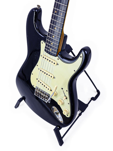 A sleek black electric guitar, the Fender L-Series Stratocaster, displayed on a stand with a minimalist white backdrop.