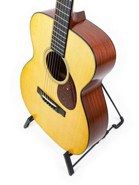 A small acoustic guitar, the Collings Julian Lage Signature OM1, elegantly displayed on a stand, showcasing its craftsmanship.

