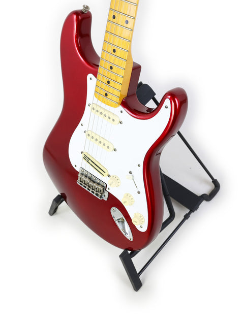 A striking red electric guitar, the Fender Vintage Hot Rod ‘57 Stratocaster, displayed on a stand with a white backdrop.
