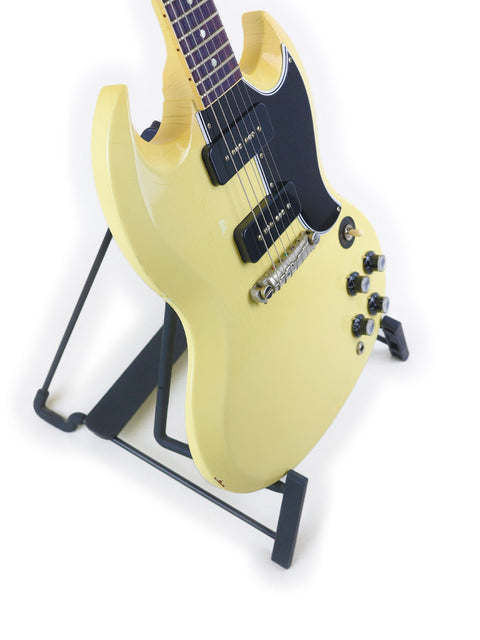 A striking yellow Gibson Custom ‘63 SG Special guitar rests on a stand, complemented by a sleek black guitar strap.