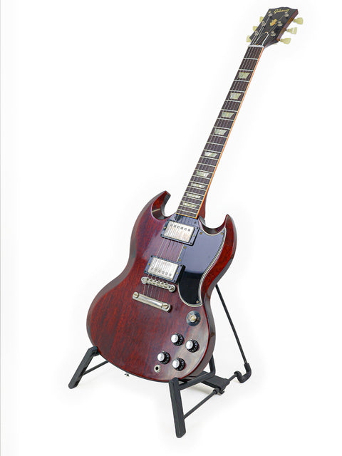  A vibrant red '64 SG Standard guitar by Gibson on a stand, set against a stark white backdrop.