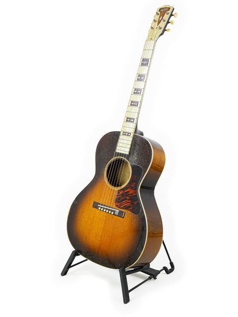 A vintage 1936 Gibson L-C 'Century of Progress' guitar elegantly positioned on a stand with a white backdrop.