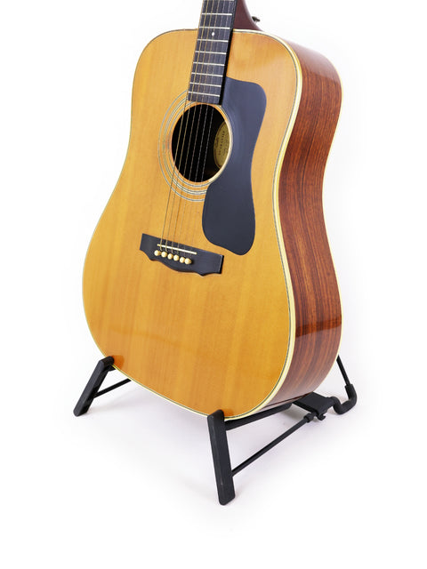 A vintage 1974 Guild D-50 Bluegrass Special guitar displayed on a stand with a plain white backdrop.
