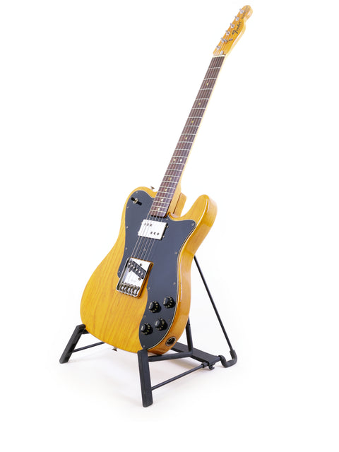 A vintage 1977 yellow Fender Telecaster Custom guitar displayed on a stand, set against a bright white backdrop.