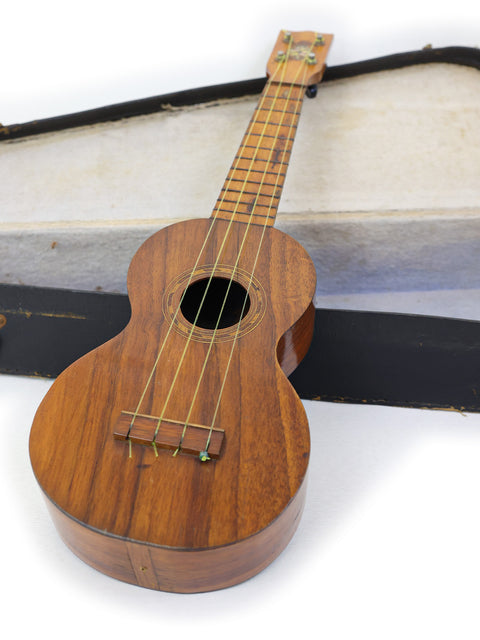  A vintage Kaai Style 2 Koa Ukulele from Hawaii (c. 1918) resting in a wooden case on a white surface.