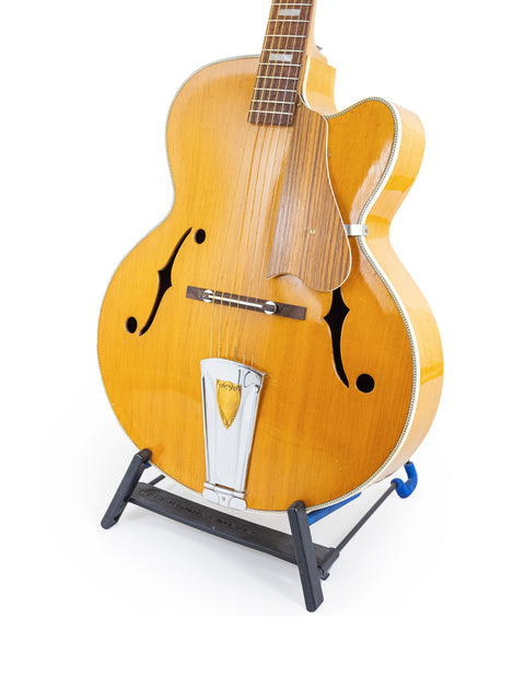 A vintage Sutton Archtop guitar from the 1970s on a stand, featuring a stark white background.
