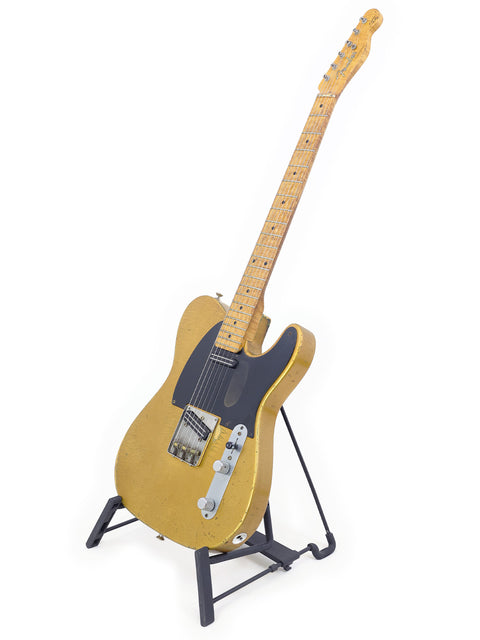 A worn gold-colored electric guitar on a black stand, against a white background.