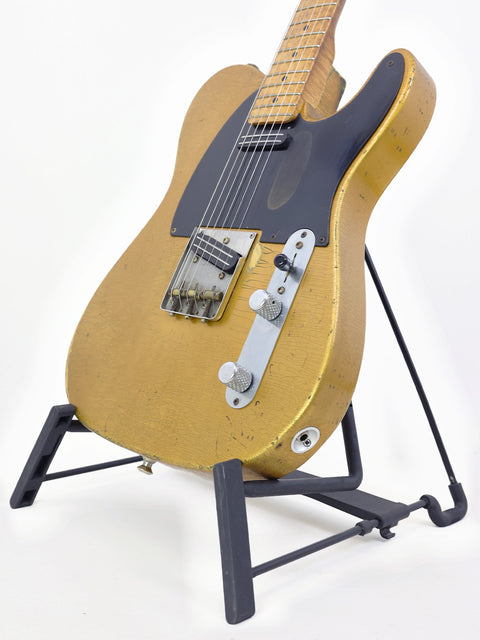 A worn gold-colored electric guitar on a black stand with a white background.