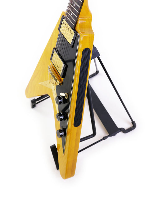 A yellow Ibanez 2387CT Rocket Roll Sr. Flying V guitar on a stand, featuring a black guitar strap.