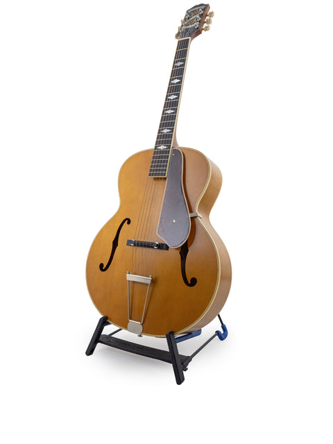 An Epiphone Masterbilt De Luxe F-Hole Archtop guitar from 2016 stands elegantly on a stand with a white backdrop.
