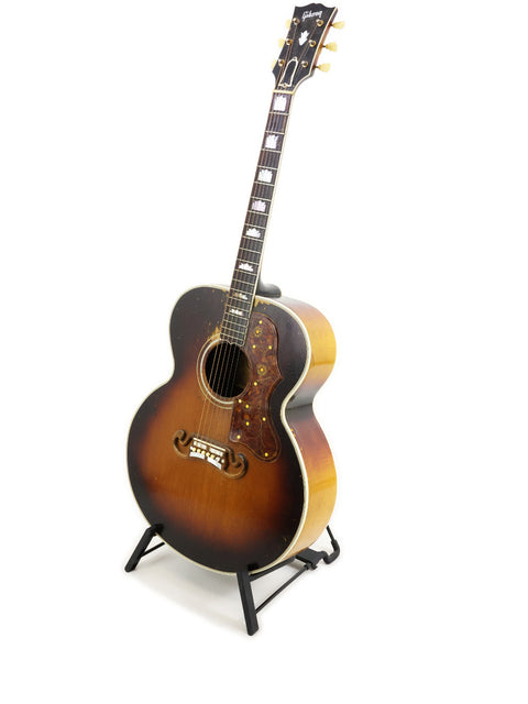 Classic 1954 Gibson J-200 guitar resting on a stand, highlighted against a simple white backdrop.