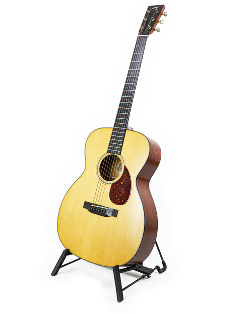 The Collings Julian Lage Signature OM1 guitar rests elegantly on a stand, contrasting with a bright white backdrop.
